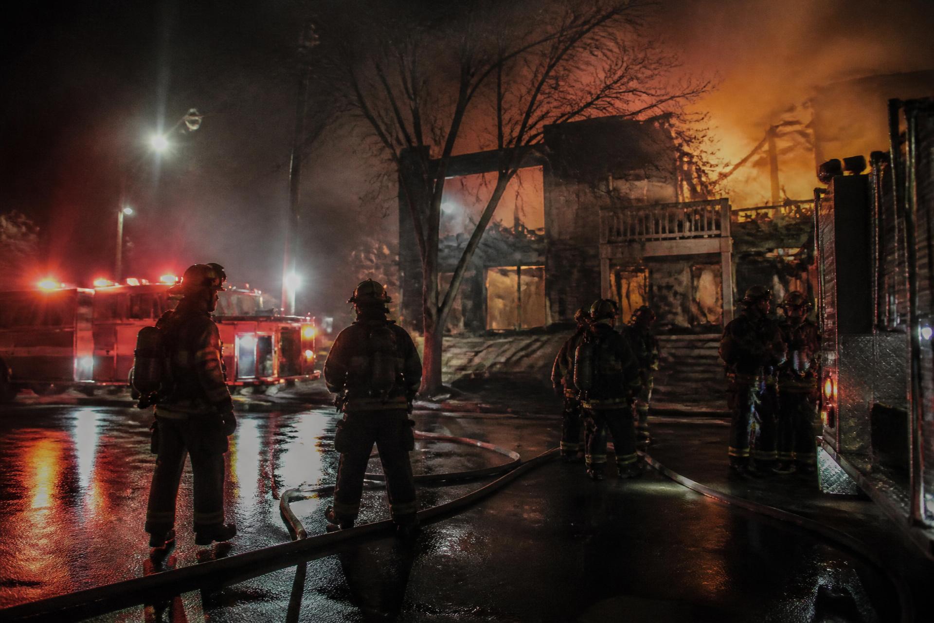 Firefighters on a wet street at night beside apparatus, fire glowing behind a building